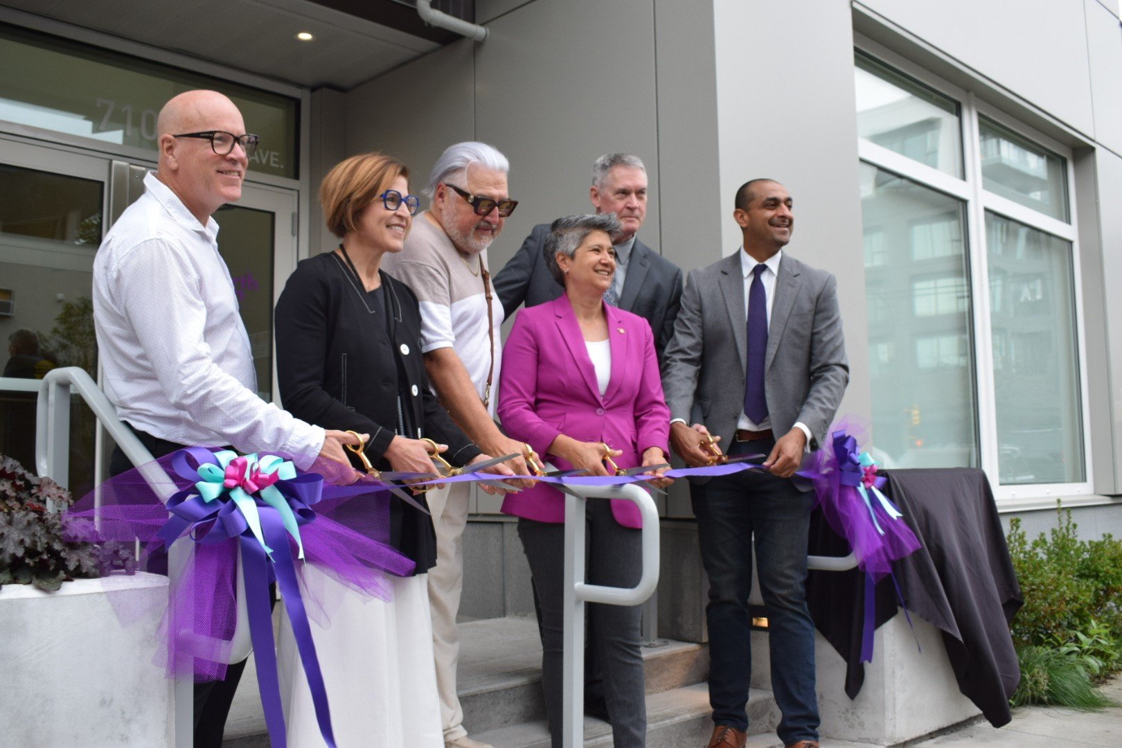 Six people stand in front of a building cutting a ceremonial ribbon with oversized scissors at a Co-operative Housing ribbon-cutting event.