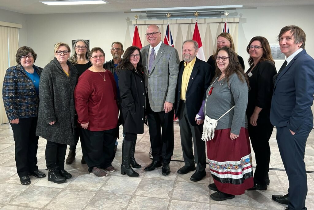 A group of twelve people stand indoors in front of several national and provincial flags, posing for a photo at a Co-operative Housing event with CHF Canada.