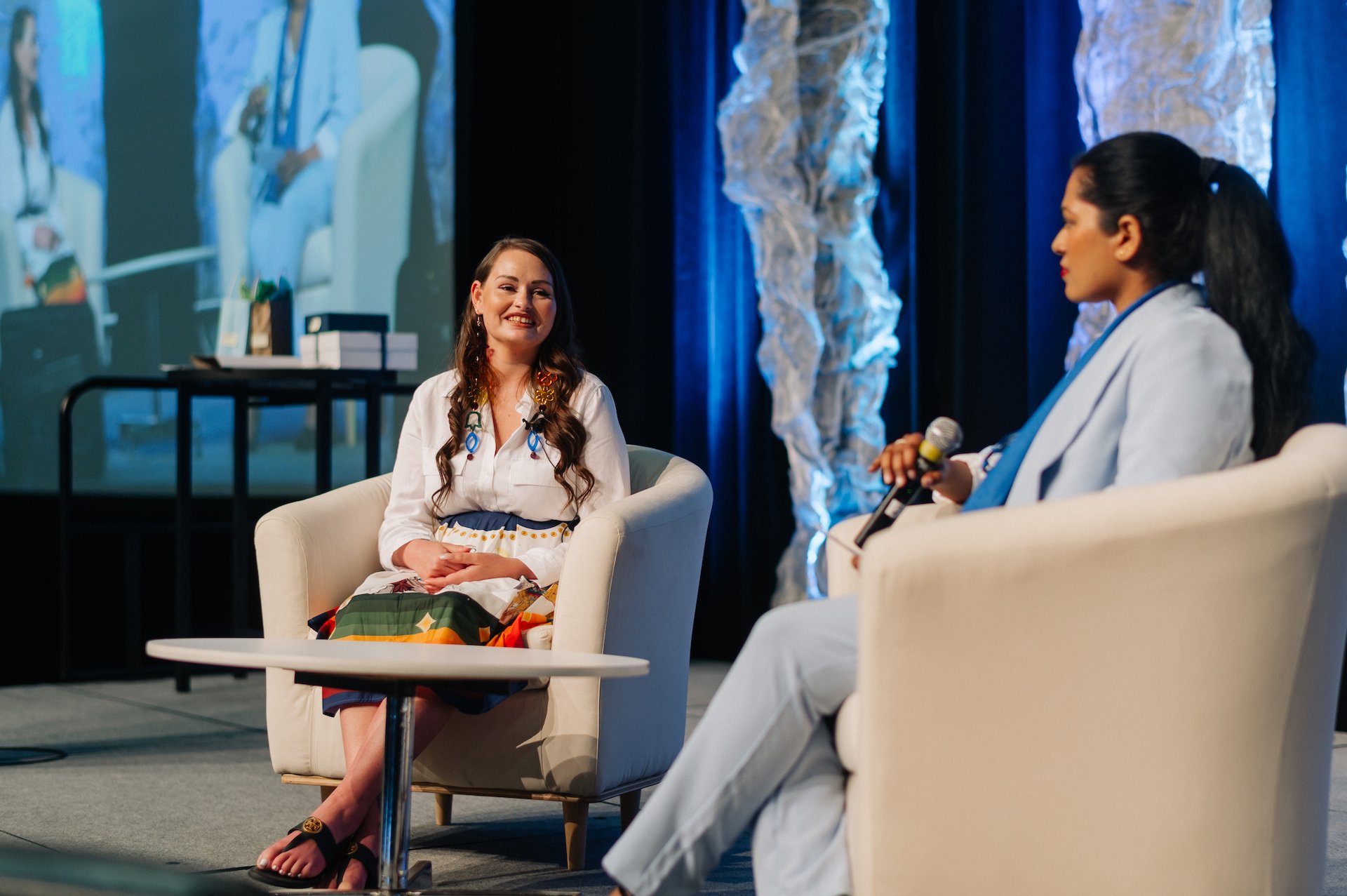 Two women sit in white chairs on a stage having a conversation about co-operative housing, with one holding a microphone. A screen in the background shows a live video feed of the CHF Canada discussion.