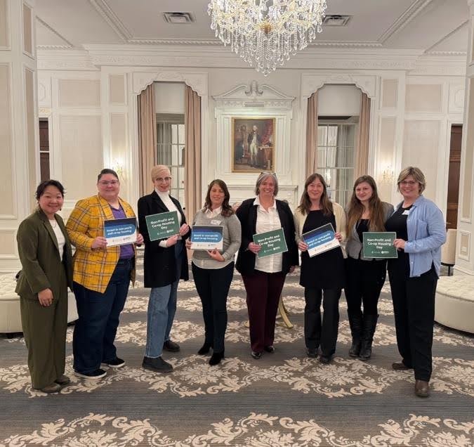 Eight women stand in a formal room, five holding green or blue signs with text supporting co-operative housing. A chandelier and a large painting are visible in the background.