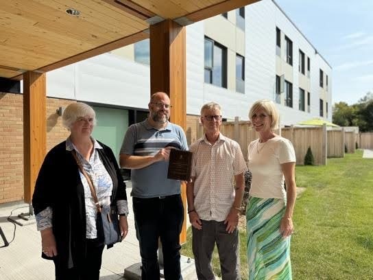 Four adults stand outside a modern building, with one person holding a plaque celebrating Co-operative Housing while the others smile for the photo.