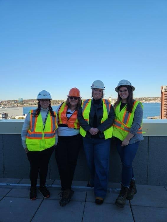Four people wearing safety vests and hard hats stand together on a rooftop, overlooking the cityscape and blue sky, showcasing their commitment to co-operative housing with CHF Canada.